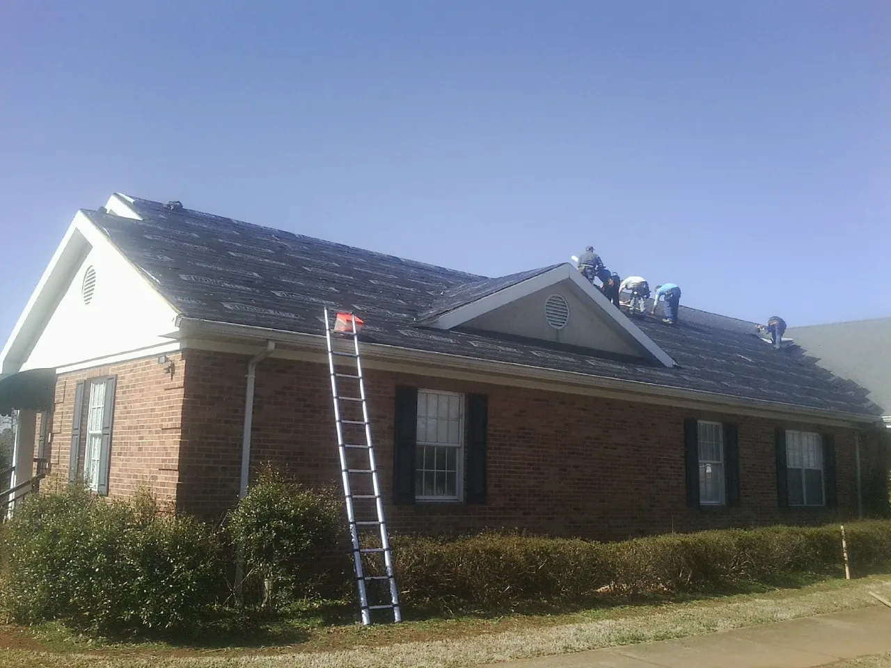 Crew tearing shingles off a brick ranch roof with a ladder set — Emergency Roof Repair work by our crew in Exira