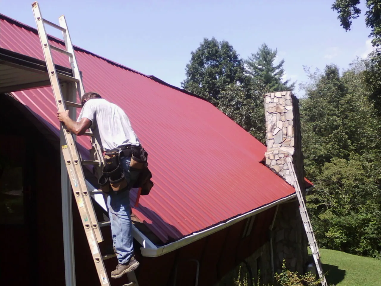 Roofer on a ladder beside a red metal roof and stone chimney — Soffit & Fascia Repair by Roofing Brothers in Exira