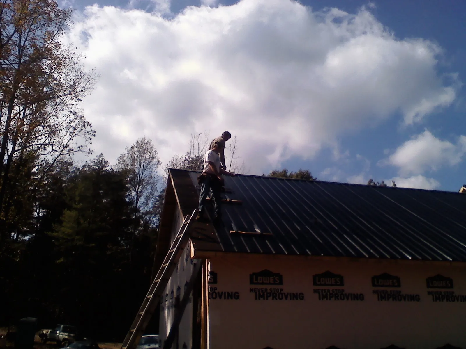 Roofer fastening dark metal panels high against a cloudy sky, from a Roof Inspection job in Exira